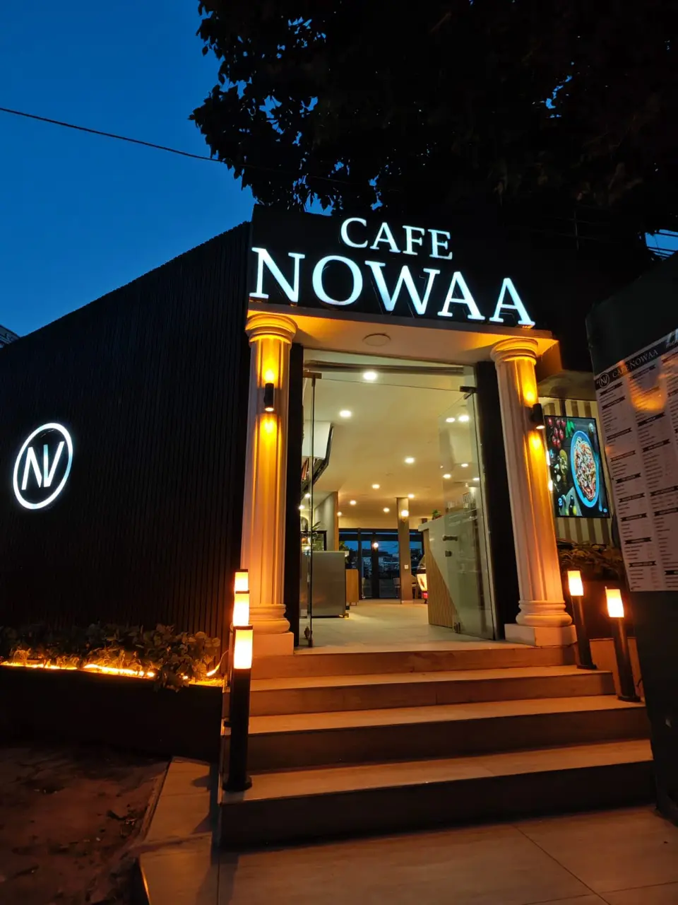 Cafe Nowaa'nın aydınlatılmış girişi ve sütunlu cephesi, akşamüstü